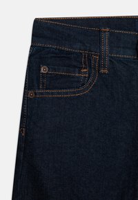 Mørkeblå denim jeans med oransje søm, en liten frontlomme og en knappdetalj nær midjen. Klassisk passform.