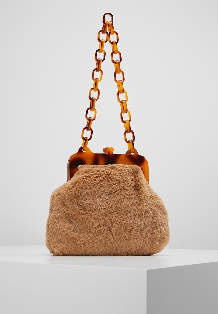 Sac à main en fausse fourrure marron avec fermoir en écaille et bandoulière à maillons épais, présenté sur un socle blanc sur fond gris.