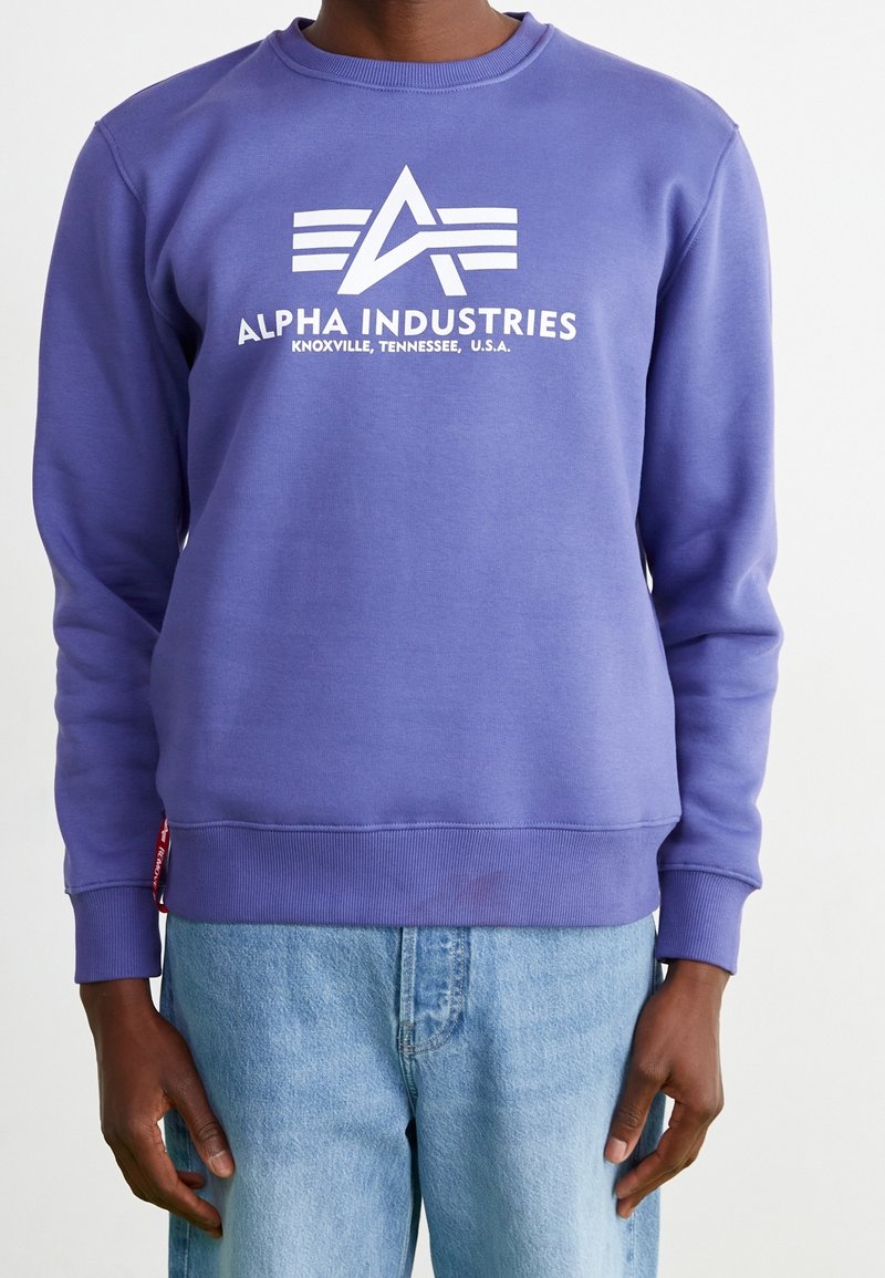 Personne portant un sweat-shirt violet avec le logo blanc "Alpha Industries" et un jean bleu clair, debout devant un fond uni.