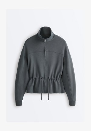 Giacca con cerniera grigio scuro con collo alto, maniche lunghe, polsini a coste e vita regolabile con coulisse, mostrata su sfondo bianco.