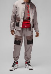 Lichtgrijze jas met zwarte gaaszakken, gecombineerd met een grijs T-shirt met rode tekst. Rode heuptas, grijze broek en zwart-witte sneakers.