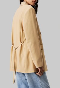 Beżowa marynarka o luźnym kroju, z tylnym wiązaniem, gładką fakturą i dwoma bocznymi kieszeniami; w zestawie z jasnoniebieskimi dżinsami.