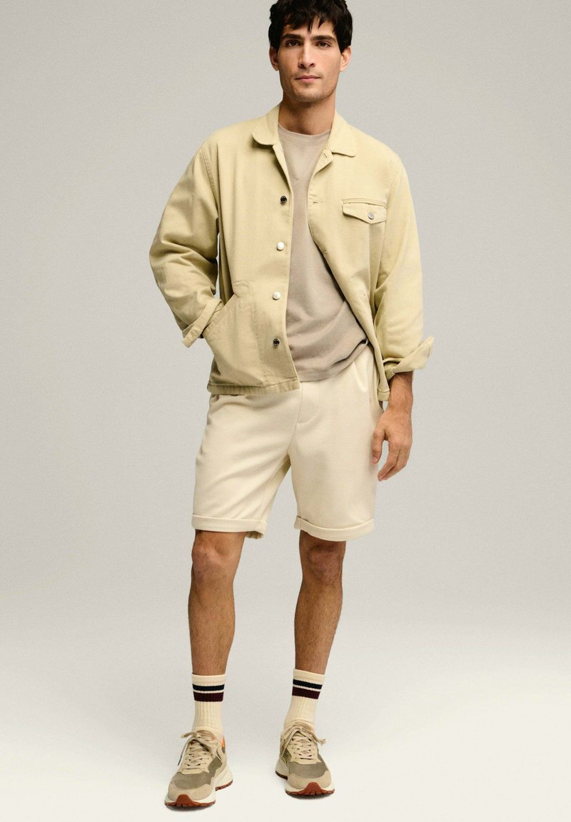 Chaqueta de algodón beige con botones, camiseta beige clara, pantalones cortos crema y zapatillas de camuflaje. Calcetines de tobillo a rayas negras y rojas.
