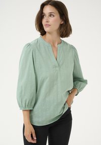 Blouse vert clair en tissu texturé, avec un col en V, des manches froncées et des motifs en relief subtils. Associée à un pantalon sombre.