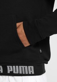 Sudadera negra de algodón con bolsillo canguro, puños acanalados y un logo blanco de "PUMA" en el dobladillo. Incluye una pequeña etiqueta del fabricante.