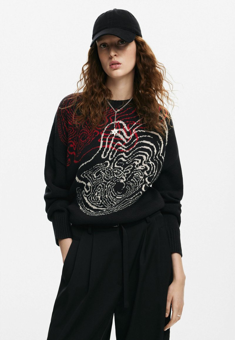 Maglione nero lavorato a maglia con intricati motivi astratti in rosso e bianco, abbinato a pantaloni neri a vita alta e un berretto nero.
