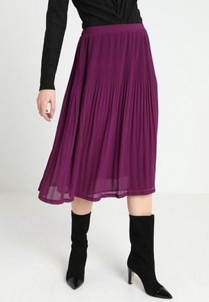 Vrouw met een paarse geplooide midirok, gecombineerd met zwarte laarzen met hakken en een zwart topje met lange mouwen, staande op een witte vloer.