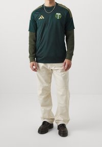 Mies, joka pitää tummaa vihreää urheilupaitaa, jossa on kultaisia logoja, oliivinvihreät pitkät hihat alla, vaaleat housut ja mustat nauhakenkiä, seisoo valkoisella lattialla.