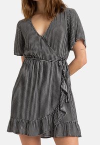 Nainen, jolla on päällä musta-valkoinen ruudullinen kietaisumekko, jossa on lyhyet hihat, V-pääntie, röyhelöhelma ja sivusiteen yksityiskohta. Mekko on näkyvissä olkapäistä reiden puoliväliin.