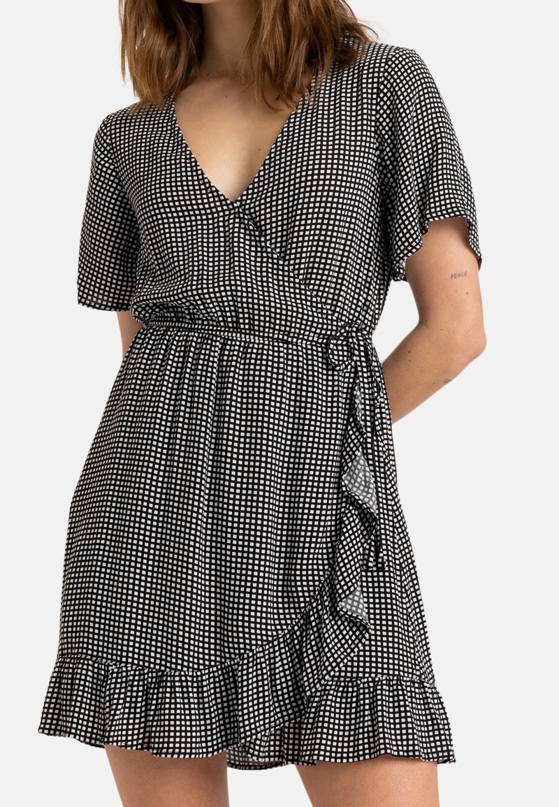 Nainen, jolla on päällä musta-valkoinen ruudullinen kietaisumekko, jossa on lyhyet hihat, V-pääntie, röyhelöhelma ja sivusiteen yksityiskohta. Mekko on näkyvissä olkapäistä reiden puoliväliin.