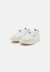 Valkoiset nahka- ja samettikengät, joissa on vaaleankermanvärinen kumipohja, tasaiset valkoiset nauhat ja pieni musta logo sivussa.