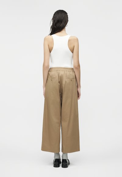 Γυναίκα με μακριά σκούρα μαλλιά που φοράει λευκό αμάνικο τοπ, φαρδιά μπεζ παντελόνια, γκρι κάλτσες και μαύρα παπούτσια, στέκεται γυρισμένη προς τα πίσω.