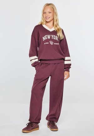 Młoda dziewczyna z długimi blond włosami, ubrana w bordowy dres z napisem New York, stoi z jedną ręką w kieszeni i uśmiecha się.