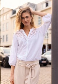 Blusa blanca de botones con detalles bordados en el cuello y las mangas, combinada con pantalones de talle alto beige que cuentan con un cinturón atado.