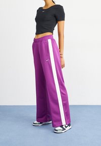 Purple wide-leg sweatpants with white side stripes and an elastic waistband, paired with a black fitted T-shirt and black, grey, and white sneakers.