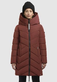 Raudona pūsta striukė su dideliu gobtuvu, zigzago raštu, priekyje užtrauktuku ir logotipo etikete. Juoda megzta kepurė, dėvima modelio galvoje.