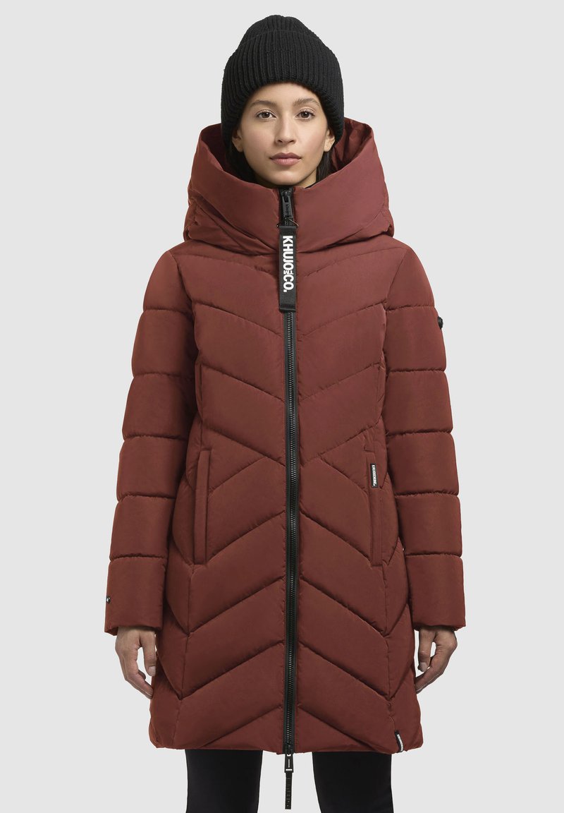 Cappotto imbottito rosso con un grande cappuccio, motivo a chevron, zip frontale e etichetta con logo. Berretto in maglia nera indossato sulla testa del modello.