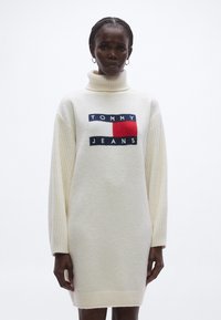 Fehér garbó ruha kötött textúrával, bordázott ujjakkal és tengerészkék és piros részletekkel díszített logófolttal.