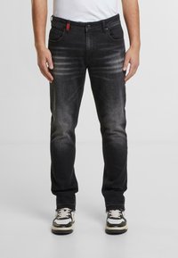 Pantalones vaqueros de mezclilla negra con un ajuste slim, diseño deslavado, ligeros desgastes y un acento rojo en la cinturilla. Combinados con zapatillas negras y blancas.