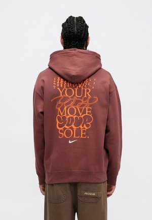 Person med fletninger, iført en brun hættetrøje med orange tekst og et hvidt Nike-logo på bagsiden, står mod en ensfarvet baggrund.