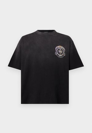 Man draagt een zwart grafisch T-shirt, zwarte spijkerbroek met riem en zwart-witte hoge sneakers, staand met één hand in de zak op witte achtergrond.