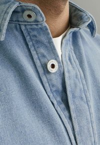 Camicia di jeans azzurro chiaro con tessuto testurizzato, dotata di colletto piegato e bottoni metallici con un logo circolare.