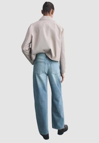 Lichtbeige oversized jas, losse blauwe jeans die vervagen naar lichter blauw, met achterzakken en minimale stikdetails. Zwarte schoenen.