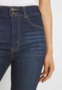 Jeans de mezclilla de tiro alto en azul oscuro con costuras visibles y dos botones en la cintura. Presentan una textura suave y un corte ajustado.