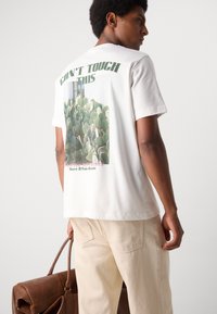 White cotton t-shirt featuring a cactus graphic on the back, with green text reading "CAN'T TOUCH THIS" above it. Casual fit, soft texture.