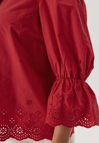 Blusa di cotone rossa con maniche a sbuffo, decorata con motivi floreali ricamati e orlo ondulato. Tessuto morbido, design leggero.