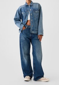 Veste en jean bleu clair avec col, poches poitrine et boutons argentés, associée à un jean large assorti et des baskets beiges.