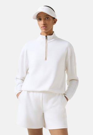 Femme portant un haut zippé blanc à manches longues, un short blanc assorti et une visière blanche, debout les mains dans les poches.