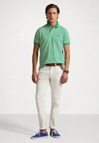 Πράσινο και λευκό ριγέ πόλο μπλουζάκι με πράσινο γιακά, σε συνδυασμό με λευκό παντελόνι και μπλε αθλητικά παπούτσια. Ορατή καφέ πλεκτή ζώνη και ρολόι.