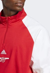 Chaqueta de quarter-zip en rojo y blanco con tejido suave, presenta un alto cuello y un logo bordado en el pecho, mangas de color contrastante.