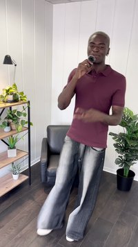 Maroon polo shirt, løse sorte og grå denim bukser, står på mørkt trægulv. Indendørs miljø med planter og en stol i baggrunden.