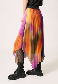 Kvinna som bär en plisserad, asymmetrisk kjol med ett flerfärgat tie-dye-mönster, svarta prickiga strumpbyxor och svarta snörade kängor.