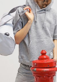 Sudadera gris con bolsillo frontal, combinada con jeans gris claro. Se lleva una bolsa blanca con correas grises. En primer plano hay un hidrante rojo.