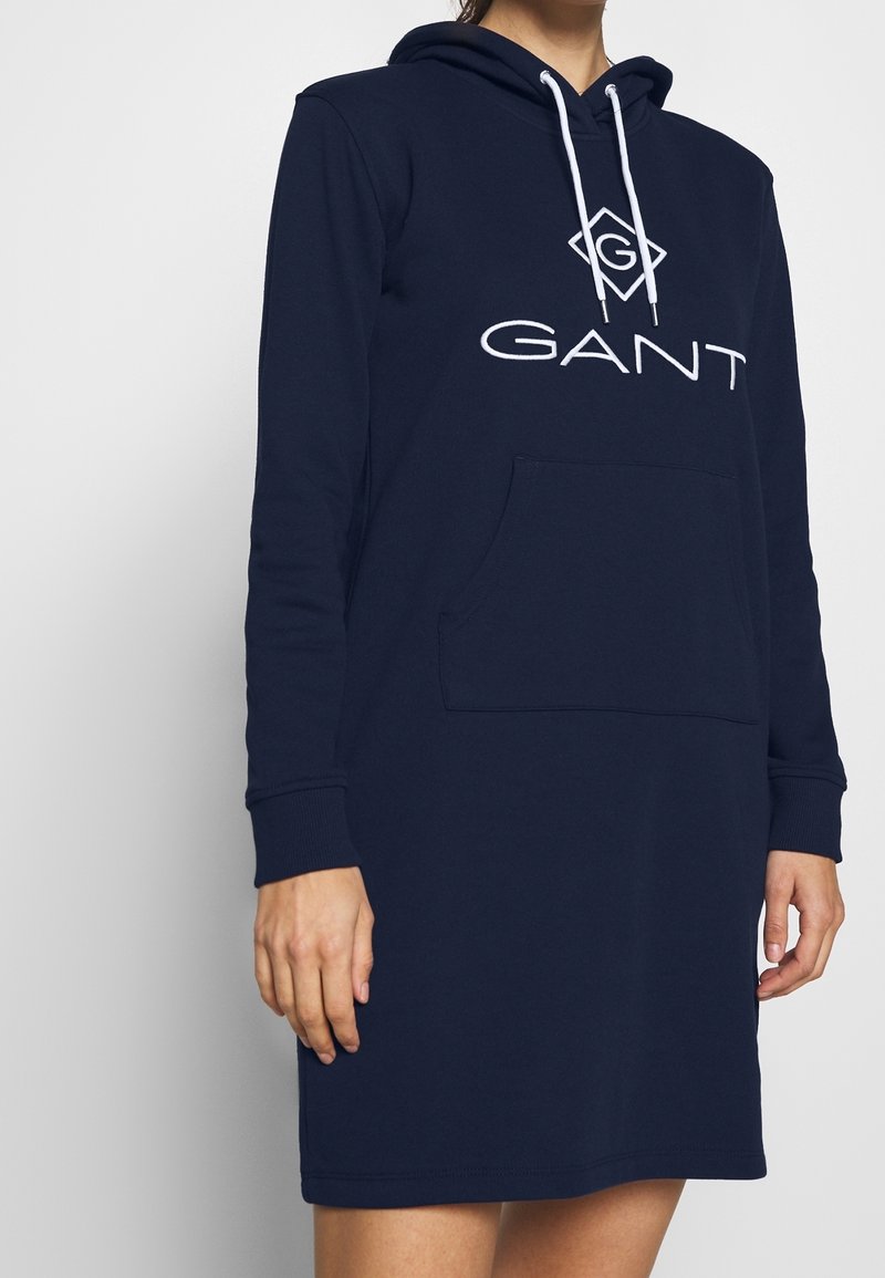 Femme portant une robe à capuche bleu marine GANT avec poche avant et cordons blancs, debout devant un fond clair uni.