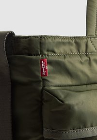 Zelena, starana torba od tkanine s trakama; ima crvenu oznaku Levi's i vidljive detalje šivanja. Glatka tekstura s oblikovanom konstrukcijom.