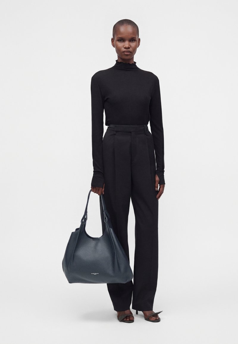 Femme portant un haut noir à manches longues et un pantalon, tenant un grand sac à main en cuir bleu foncé avec des sandales à talons hauts sur fond blanc.