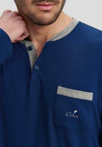 Camisa azul de manga larga con un cuello henley gris, dos botones azules y un bolsillo gris con un logotipo bordado en blanco. Tela de algodón suave.