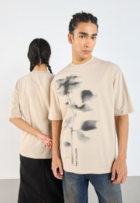 Camiseta oversized beige con un gráfico floral oscuro y la palabra "NOCTIFLORA" impresa en el lado inferior. Material de algodón; mangas cortas.