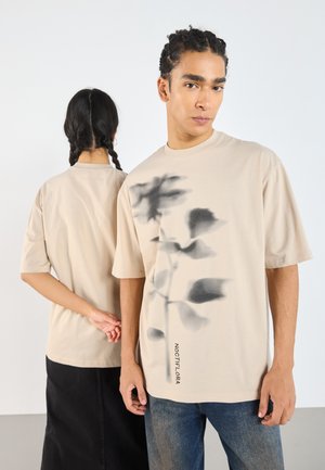 Beige oversized t-shirt med en mörk blommig grafik och ordet "NOCTIFLORA" tryckt längs den nedre sidan. Material av bomull; korta ärmar.