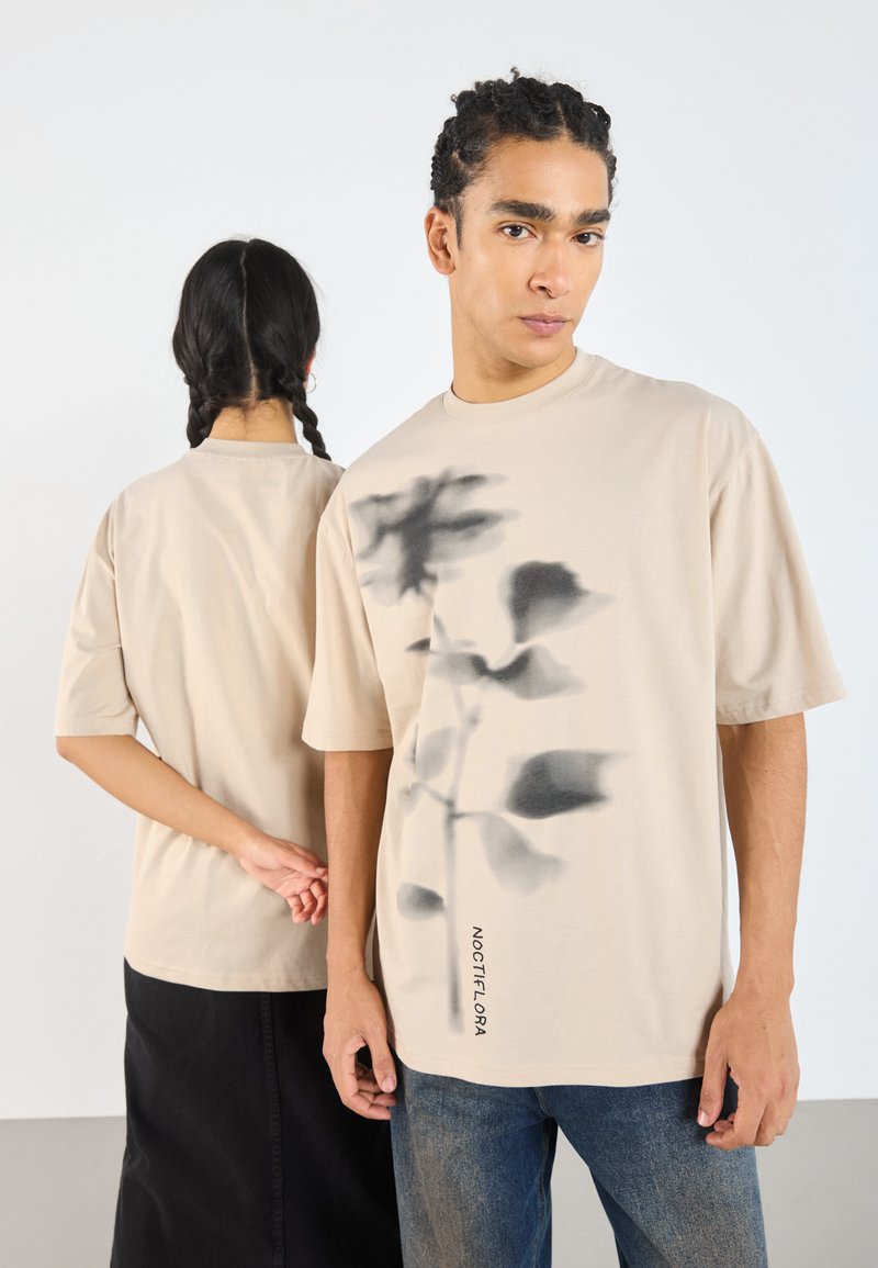 Camiseta oversized beige con un gráfico floral oscuro y la palabra "NOCTIFLORA" impresa en el lado inferior. Material de algodón; mangas cortas.