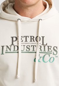 Cremfarbener Hoodie aus Baumwolle mit Kapuze und Kordelzug. Auf der Vorderseite prangt in fettem Schwarz und Grün der Text "PETROL INDUSTRIES & Co."