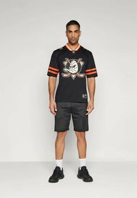 Črna hokejska majica z oranžnimi akcenti in logotipom na prsih, v kombinaciji s črnimi mrežastimi šortami in črnimi supergami. Vidne so navadne bele nogavice.