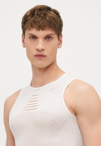Top sans manches blanc avec un design texturé comportant des motifs en maille circulaires et triangulaires, et un col lisse. Le tissu semble extensible.