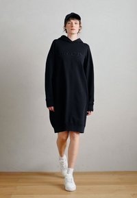 Čierne oversize mikinové šaty s textúrovaným logom na hrudi, s dlhými rukávmi, kapucňou a rebrovanými manžetami, doplnené o biele tenisky.