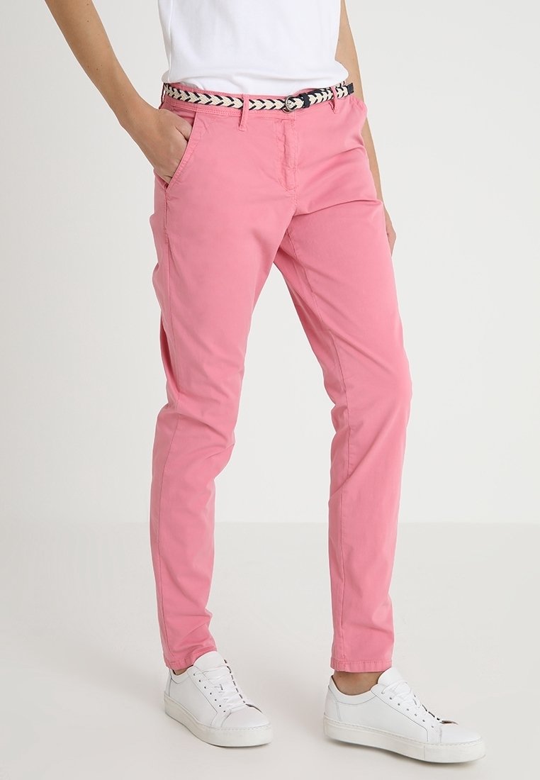Personne portant un pantalon rose slim avec poches latérales, une ceinture tressée, des baskets blanches et une chemise blanche, debout devant un fond uni.