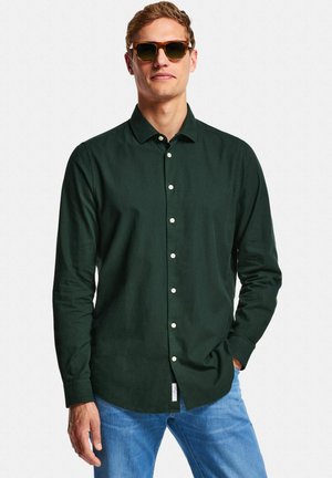 Man draagt een donkergroen overhemd met knopen, blauwe spijkerbroek en een zonnebril, staat met één hand in de zak tegen een witte achtergrond.